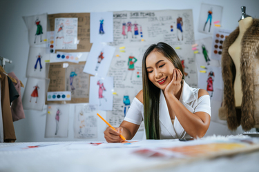 Young Asian designer female drawing fashion sketch in workshop. Beautiful clothes sketch picture on board behind her
