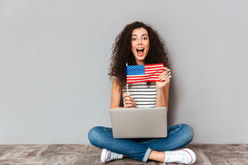 Gorgeous female with beautiful smile sitting in lotus pose with silver computer on legs, demonstrating american flag on camera over grey wall